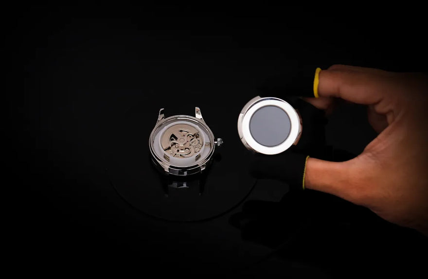Hand holding a watch lens over a watch mechanism on a dark background