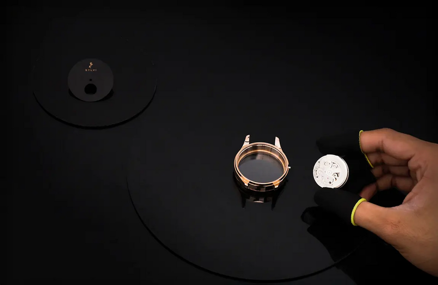 Watch components on a black surface with a hand holding one of them.