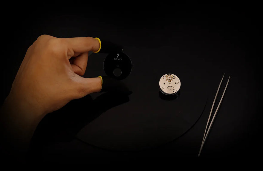 Hand holding a small black device on a dark surface with a coin and tweezers.